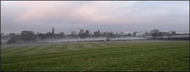 View across misty fields with trees and church tower in the distance.